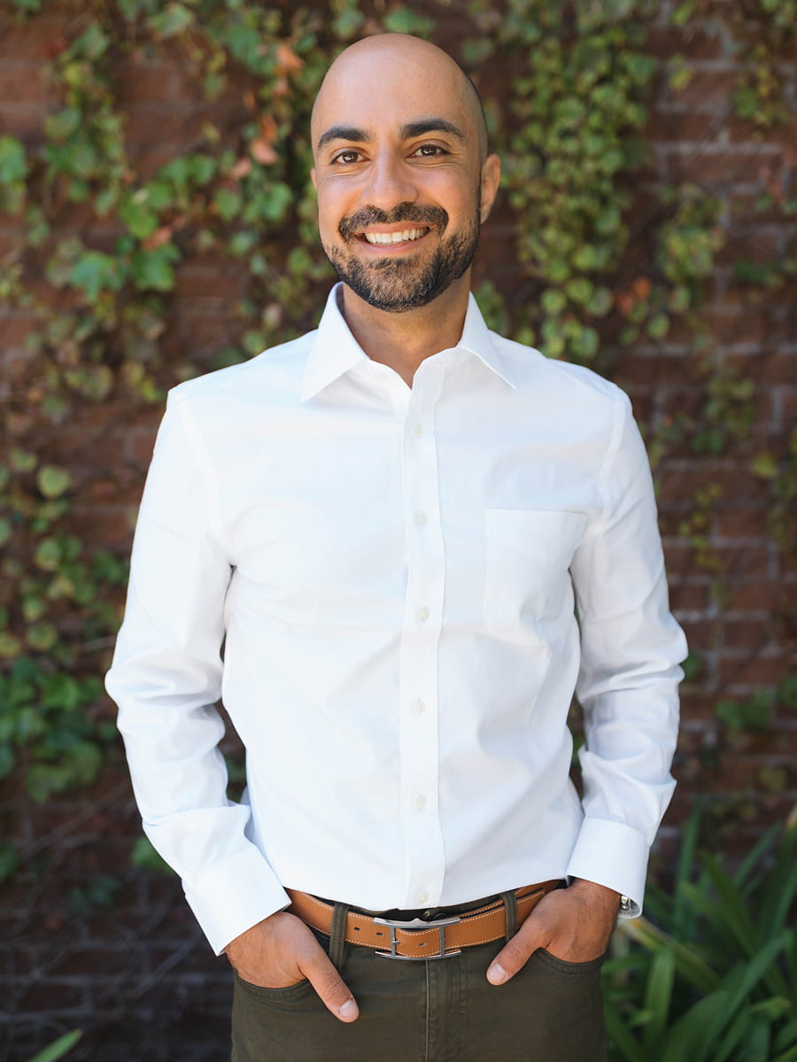 A person wearing a white button-up shirt and brown belt stands outdoors in front of a brick wall with green ivy, smiling with hands in their pockets.