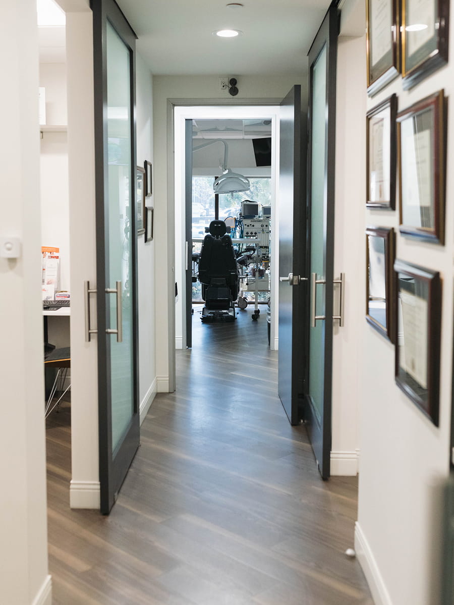 A hallway with glass doors leads to a dental office where a patient sits in a dental chair. Medical equipment is visible, with framed certificates on the wall to the right.