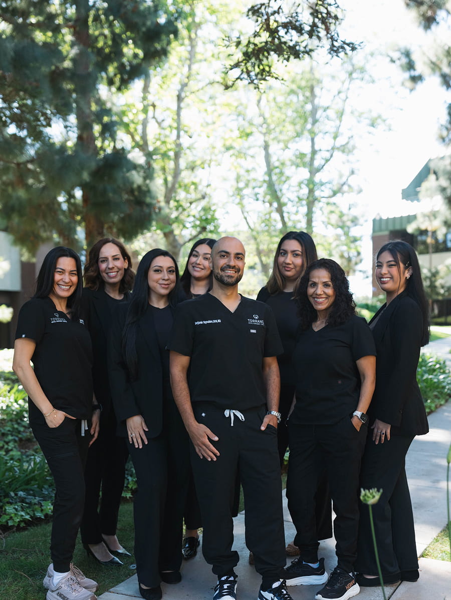 Eight people wearing black clothing stand together outdoors on a path, surrounded by greenery and trees, smiling at the camera.