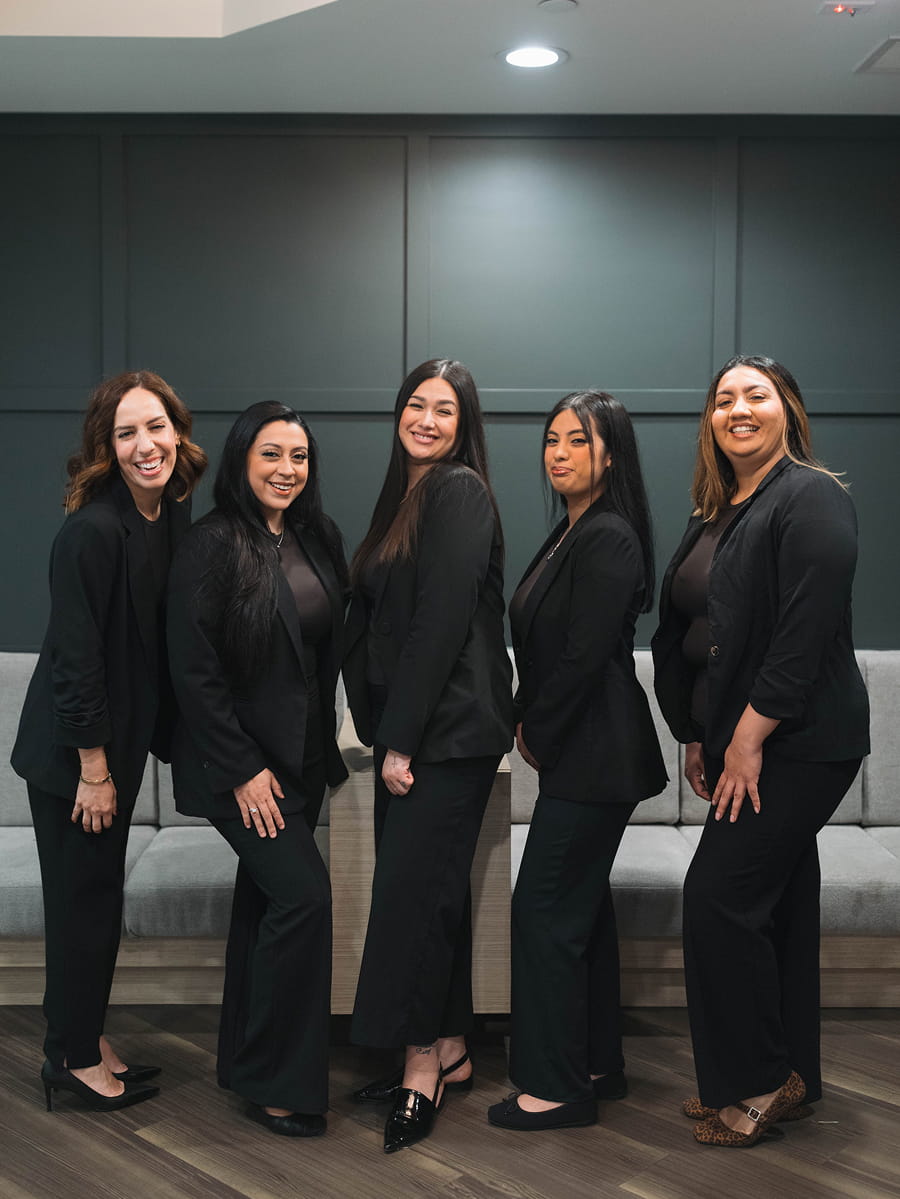 Five women in black business attire stand and smile together in an office setting with a gray bench and dark green wall in the background.