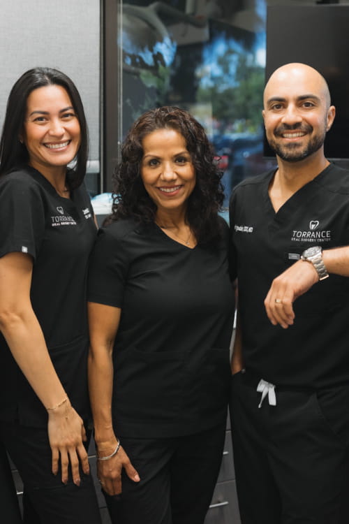 Three dental professionals in black scrubs stand together and smile in a dental office setting.